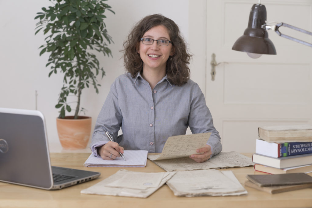 A white woman with curly brown hair and glasses is sitting at a wooden desk and smiles right at the camera. A noteblock and several old documents are lying in front of here. There are also some books and a laptop on the table.