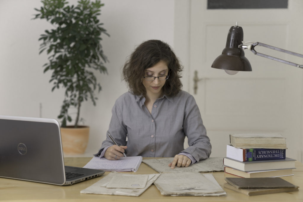 Eine junge Frau sitzt an einem Schreibtisch, auf dem sich verschiedene Unterlagen, Bücher und ein Laptop befinden. Ihr Blick ist auf ein altes Dokument gerichtet.