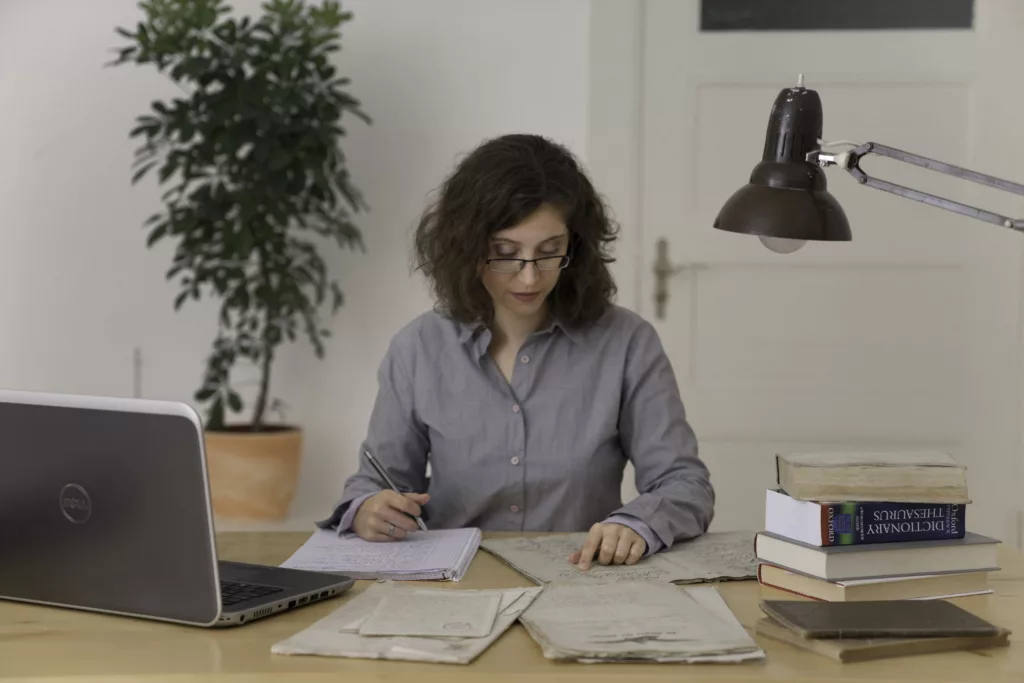 Eine weiße Frau mit braunen Locken und Brille sitzt an einem Schreibtisch und arbeitet konzentriert. Vor ihr liegen ein Schreibblock und einige alte Dokumente, außerdem ein Laptop und mehrere Bücher.